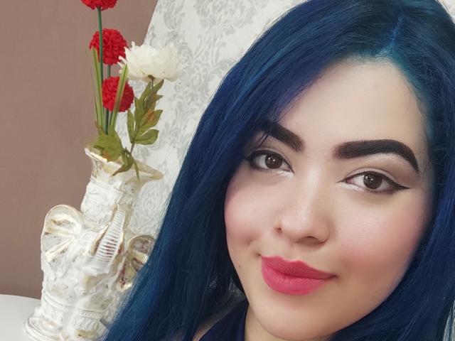 KimLafayette has blue hair, brown eyes, and pink lipstick. She's smiling with red and white flowers in a white vase with a sculpted figure on the left.