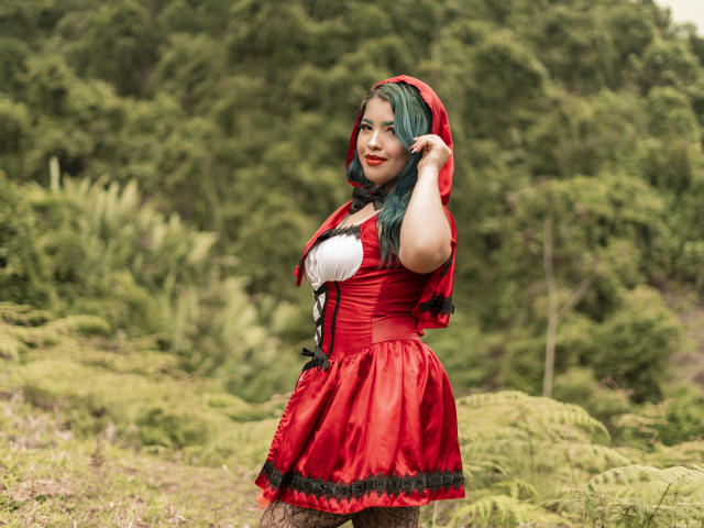 KimLafayette in a red Little Red Riding Hood costume with a white lace-up bust, green hair, red hood, and black lace trim, standing in a forest, touching her hair.