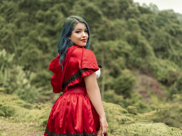 KimLafayette with blue hair, red dress, black lace trim, and a red bow, looking over her shoulder with a slight smile, standing in a green forest.
