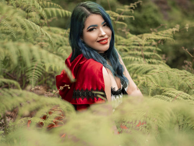 KimLafayette with blue hair, red cloak, and black lace dress, smiling with red lipstick, surrounded by green ferns, showing her tits and cute face.