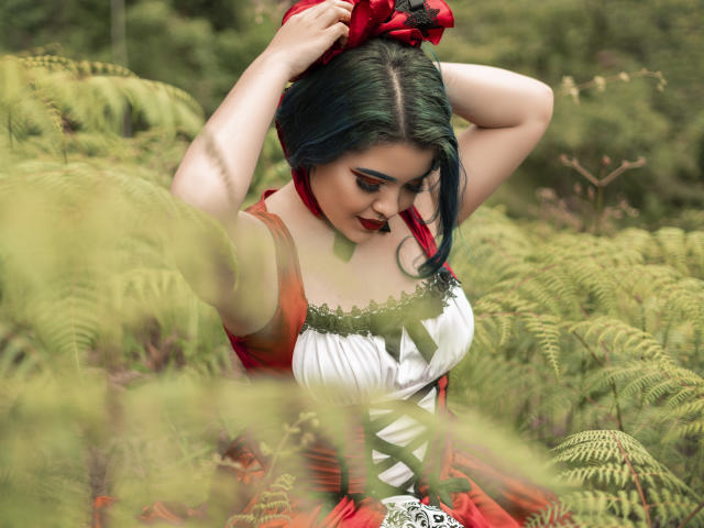 KimLafayette, avec ses cheveux bleus, sa robe rouge et son corsage blanc, ajuste un nœud rouge, regarde vers le bas, entourée de fougères vertes, montrant sa poitrine et ses lèvres rouges..