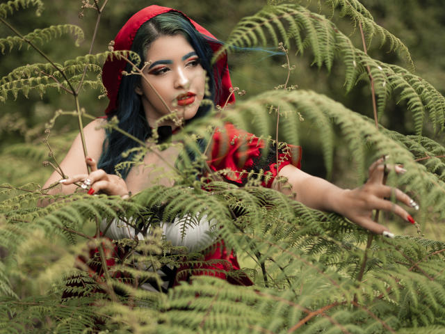 KimLafayette, com cabelo verde escuro, capuz vermelho e vestido vermelho, cercada por samambaias verdes, tem batom vermelho, sombra azul e está tocando as samambaias com unhas pintadas de vermelho..