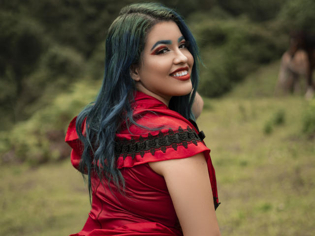 KimLafayette con i capelli blu-verdi, un vestito rosso, bordi in pizzo nero, sorride alla fotocamera, mostra la schiena, sexy, carina e attraente, all'aperto, sfondo di foresta, luce soffusa, aspetto naturale..