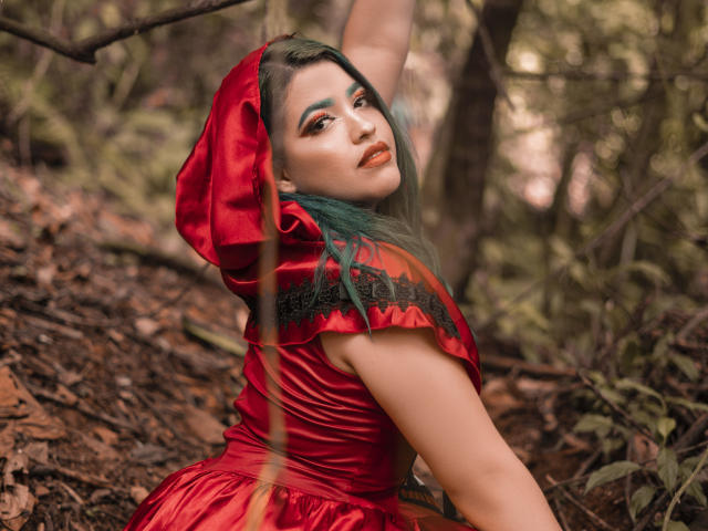 KimLafayette in un abito di seta rossa con un cappuccio, capelli verdi e un trucco audace, che si guarda indietro in una foresta, con un braccio alzato, mostrando il seno e i glutei..