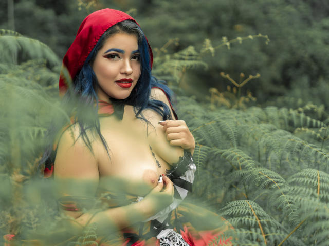 KimLafayette con i capelli blu, un cappuccio rosso e labbra rosse, che mostra il seno, tenendosi il capezzolo, in una foresta verde con felci, che guarda la fotocamera con un leggero sorriso..