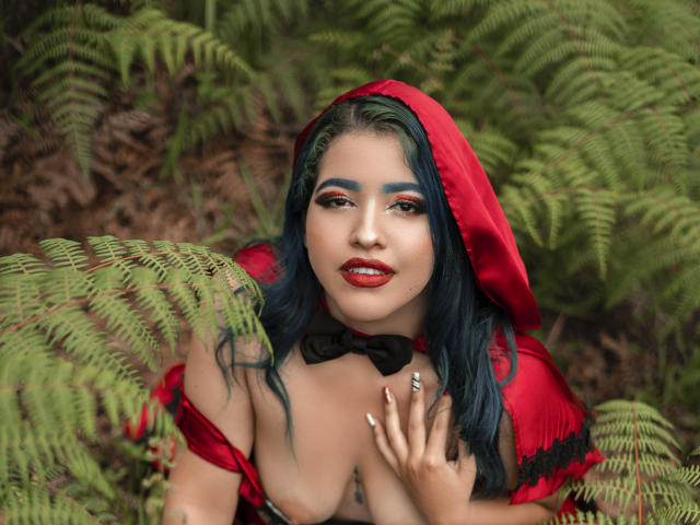 KimLafayette with blue hair, red hood, black bowtie, and red lipstick, has her hand on her cleavage, surrounded by ferns, with green eyes and red eyeshadow.