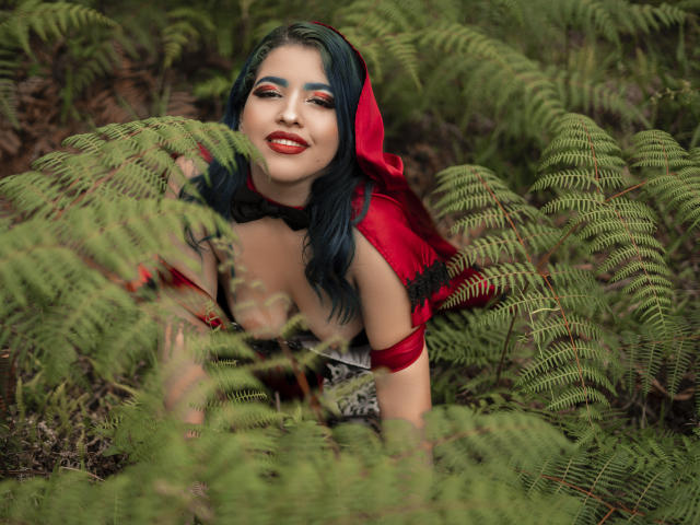 KimLafayette, con i capelli blu, gli occhi rossi e il rossetto rosso, è accovacciata tra felci verdi, indossa un cappuccio rosso e un top nero e rosso che mette in mostra il seno. Sorride..