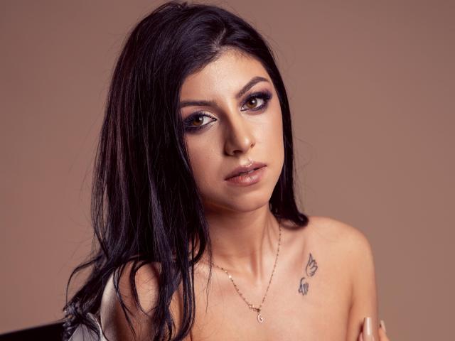 KateSparks, con cabello oscuro y húmedo, ojos seductores y labios marrones brillantes, sin ropa, mostrando pequeños tatuajes cerca de sus pechos, usando un collar dorado, fondo marrón..