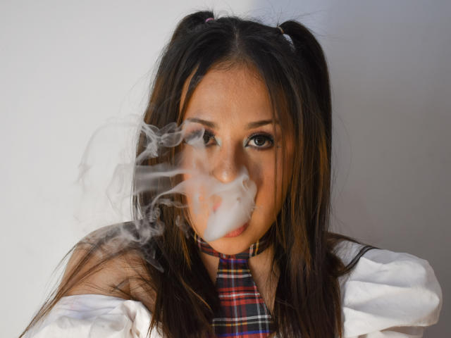 Katie Woolf, avec de longs cheveux bruns attachés en couettes, du tabac près de son visage, portant une cravate à carreaux et une chemise blanche, regardant directement l'appareil photo avec une expression sérieuse..