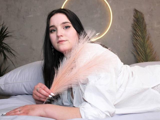 Lianna, com cabelo preto, camisa branca, segurando grama-rosa, deitada na cama, com uma luz de anel neon, uma planta verde e uma planta marrom ao fundo. Seios não visíveis..