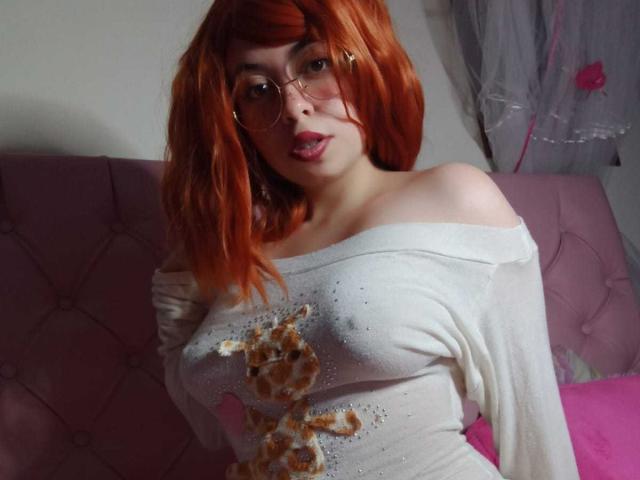 SummerTy, con cabello rojo, gafas y una blusa blanca con un hombro descubierto que muestra pequeños senos, un estampado de jirafa y un cojín rosa, sacando la lengua..