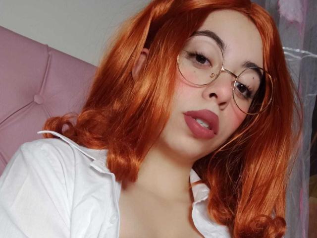 SummerTy, con cabello rojo, usando gafas redondas y una camisa blanca, mostrando sus labios rosados y su piel clara. Sus senos son ligeramente visibles debajo de la camisa..