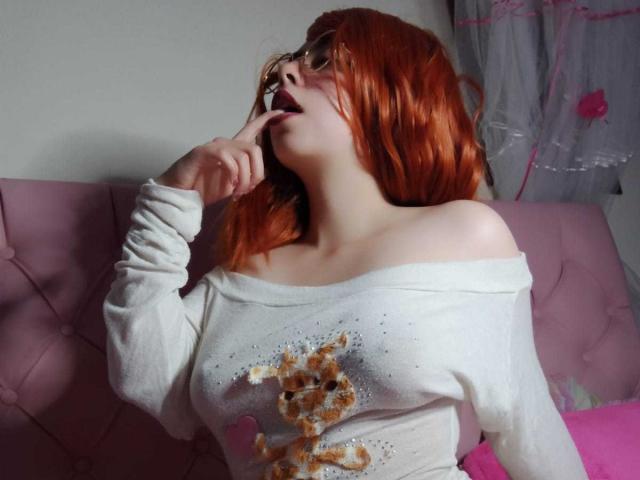 SummerTy, con i capelli rossi, indossa un maglione bianco con una spalla scoperta e ha un dito in bocca, seduta su un divano rosa, mostrando il seno e una parte superiore scintillante con un motivo marrone..