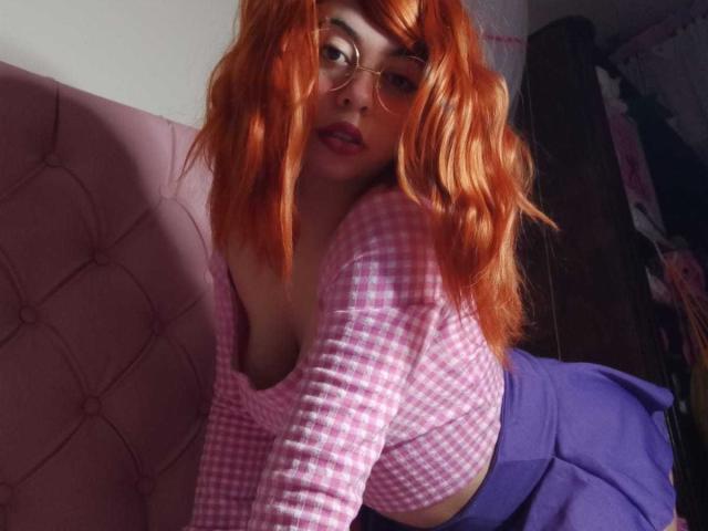 SummerTy met oranje, golvende haren, een bril, een roze geruit top, waarbij haar borsten zichtbaar zijn, leunt tegen een roze gestoffeerde bank, draagt een blauwe rok en kijkt naar de camera met een licht geopende mond..