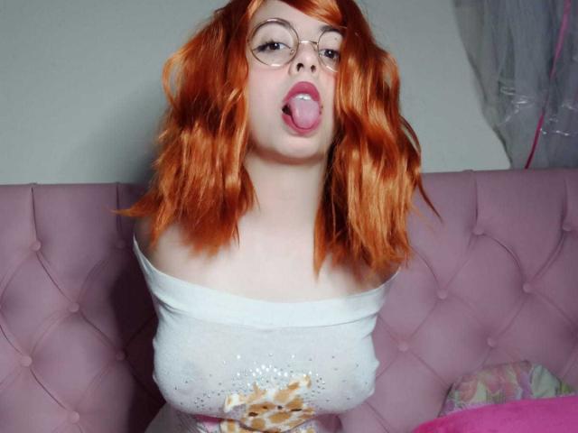 SummerTy, con cabello ondulado naranja, gafas y la lengua afuera, vestida con una blusa blanca con un hombro descubierto, mostrando sus pechos, sentada en un sofá rosa con adornos..