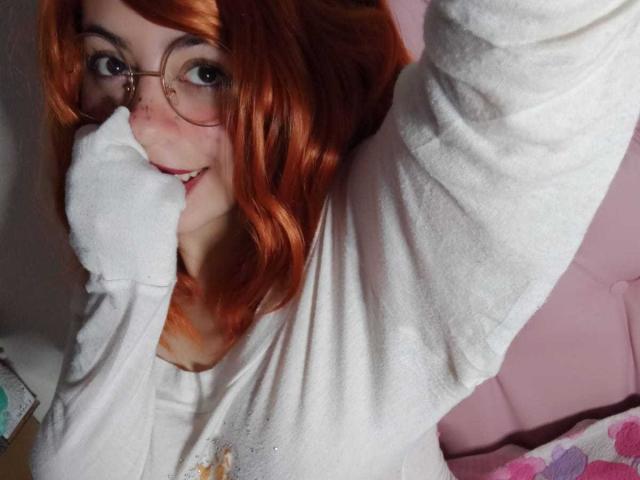 SummerTy with red hair, glasses, and white sweater, covering her nose and smiling, her arm raised, showing her cute freckled face and slightly visible tits.