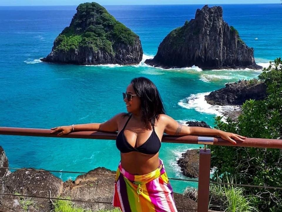 MariFun met grote borsten, een zwarte bikini top, een kleurrijke sarong, zonnebril, armen op de reling, staand bij een rotsachtige klif, blauwe oceaan, twee groene rotsachtige eilanden op de achtergrond, een zonnige dag..