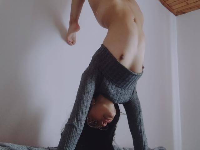 EmilyHottest staat op zijn kop, draagt grijze geribbelde broek en haar kleine borsten en platte billen zijn zichtbaar. Ze heeft een bril en zwart haar, met haar hoofd tussen haar benen..