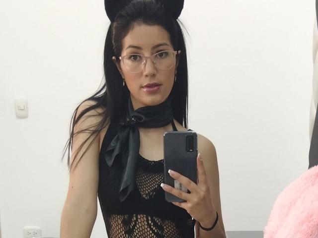 EmilyHottest with black cat ears, glasses, and black fishnet top, holding phone, showing bare shoulders and chest, pink lipstick, black hair, black ribbon, pink cushion in background.