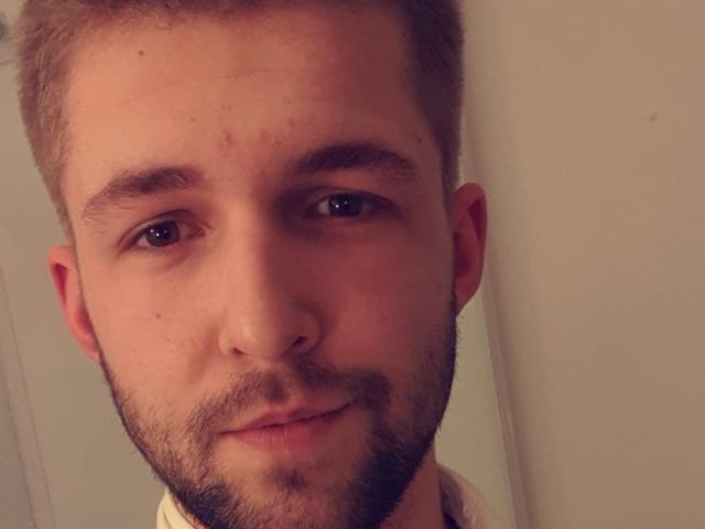 HomTeo's face with short brown hair, light brown eyes, slight beard, small nose, and subtle smile. Small scar above left eyebrow. Warm, slightly blurry selfie.