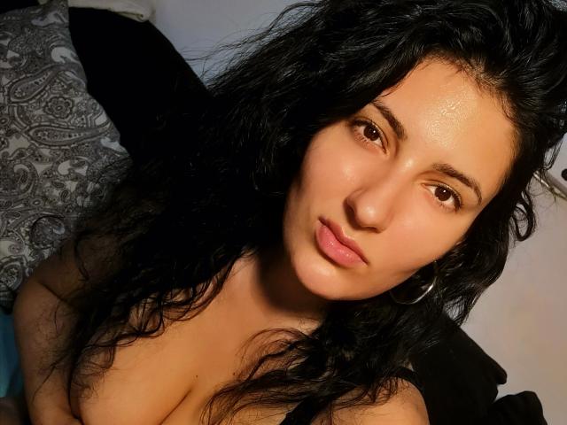 AddictedToUTS has wavy black hair, brown eyes, and pink lips. She's topless with visible cleavage, wearing a silver hoop earring. Her skin is tan and she looks relaxed.