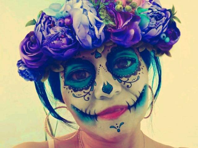 SweetieOnline hat ein bemaltes Gesicht mit blauem und grünem Augen-Make-up, einem violetten Blumenkranz und einem weißen Totenkopf-Design. Blaue Lippen und Nase, violette Rosen und blaues Haar..