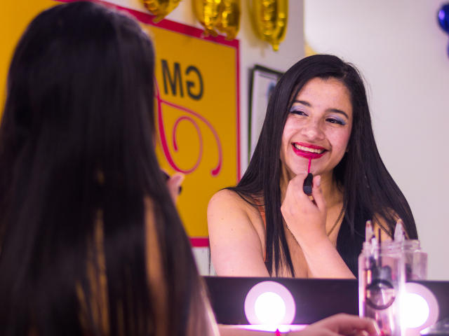 JanaSweet with black hair, smiling, holding a red lipstick to her shiny pink lips, standing in front of a mirror with makeup lights, topless, in a brightly lit room.