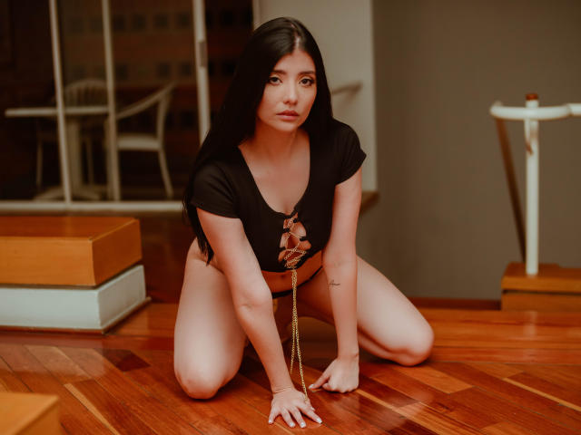 CatySuare, agenouillée sur un plancher en bois, portant un crop top noir avec découpes, laissant entrevoir de petites seins, des sous-vêtements noirs, tenant une chaîne, avec de longs cheveux noirs, des yeux marrons, et une expression sérieuse..