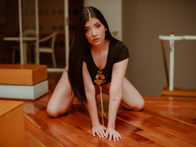 CatySuare is on her knees, arms forward, wearing a black top with a red ribbon, exposing her tits. She has long black hair, a tattoo on her right arm.