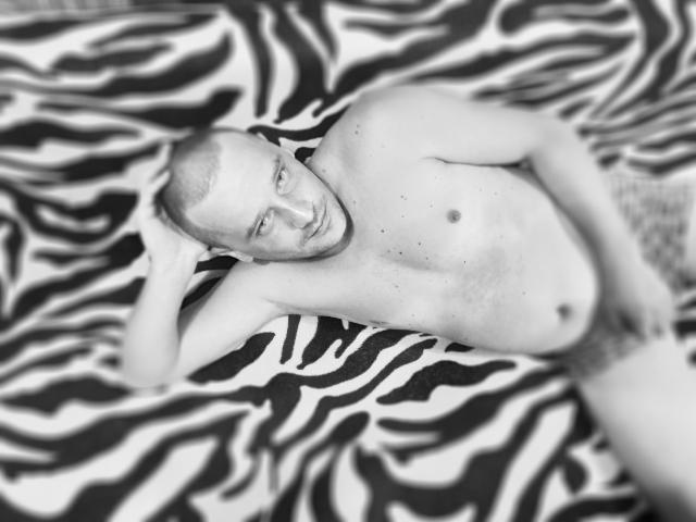 Schwarz-weißes Foto von DanielCool, der nackt auf einem bett mit Zebra-Muster liegt, mit rasiertem Kopf, Bart und einer Hand im Haar, wobei ein schlaffer, unbeschnittener Penis zu sehen ist..