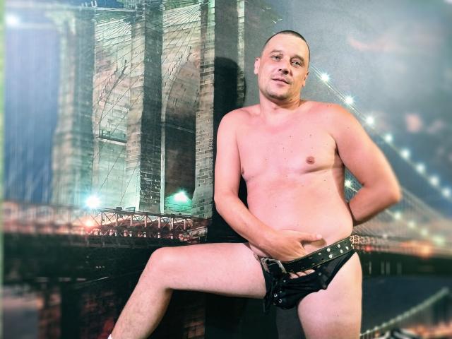 DanielCool, sem camisa e com a cabeça raspada, vestindo uma sunga de couro preta, com a mão cobrindo sua virilha, em pé em frente a um cenário de ponte, olhando ligeiramente para o lado, com uma expressão confiante..