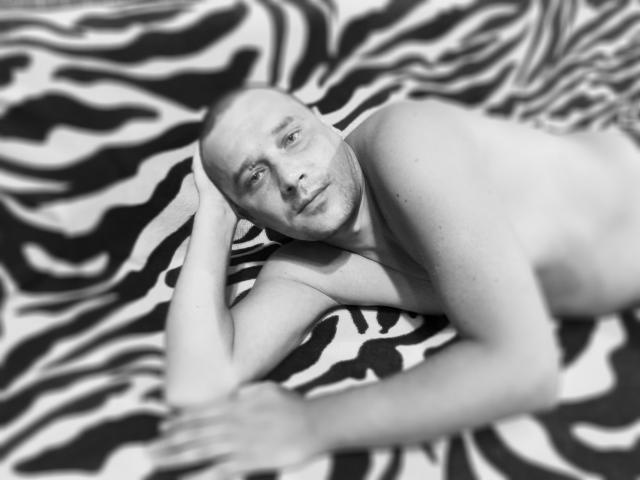 Foto in bianco e nero di DanielCool, calvo, sdraiato su una coperta a motivi zebrati, con la testa appoggiata al braccio, che guarda la fotocamera, senza camicia, con un leggero sorriso, illuminazione soffusa, rilassato, leggermente sfocata..
