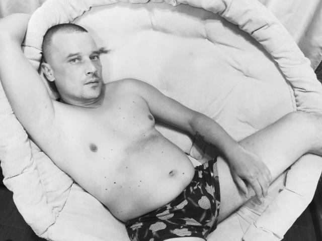 Photo en noir et blanc de DanielCool, torse nu, avec la tête rasée, allongé dans un fauteuil rond rembourré, portant un slip noir à motifs, une main sur l'aine, regardant l'appareil photo..