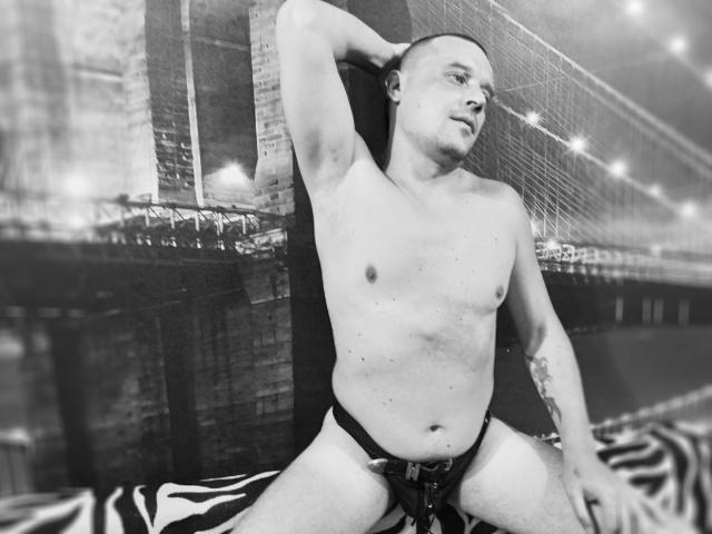 Foto em preto e branco de DanielCool, sem camisa, usando uma sunga, sentado com um braço atrás da cabeça, tatuagem no antebraço esquerdo, cobertor com padrão de zebra, olhando para o lado..