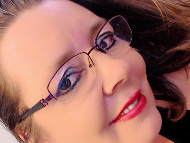 A selfie of MilfXX with glasses, blue eyes, red lipstick, and brown hair, showing a slightly winking smile. Her tits are partially visible. Her face is centered.