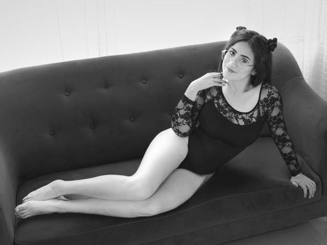 Charlotte Baker está deitada em um sofá escuro e acolchoado, vestindo um collant de renda preta que revela suas nádegas e pernas, usando adoráveis tiaras com orelhas de gato, sorrindo, em uma foto em preto e branco..