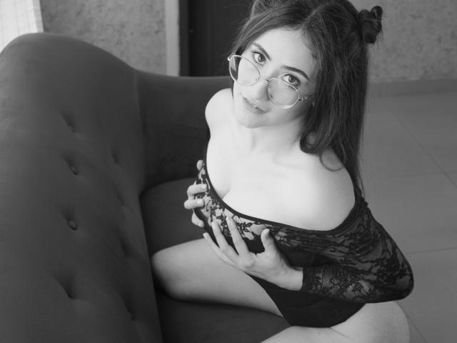 CharlotteBaker with glasses, black lace off-shoulder top, and bare tits, sitting on a couch, holding her tits with her hands, looking up with a slight smile.