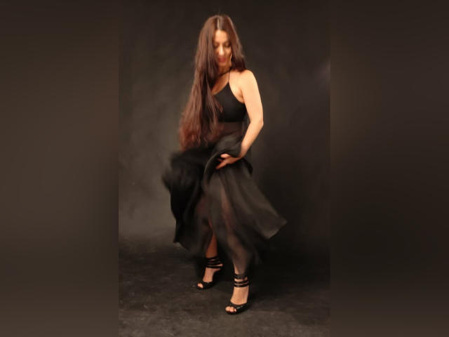 GinaONeon with long brown hair, black one-shoulder dress, and black strappy heels, slightly bent over, holding dress up, showing her ass, in a dark, smoky studio.
