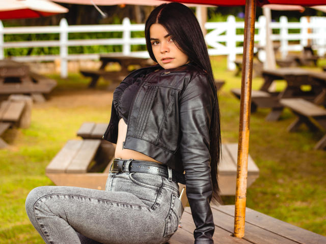 BrithneyLee, com cabelos longos e pretos, jaqueta de couro preta e calças jeans cinzas, sentada em uma mesa de madeira, mostrando suas nádegas, sob um guarda-chuva vermelho, com uma cerca branca ao fundo..
