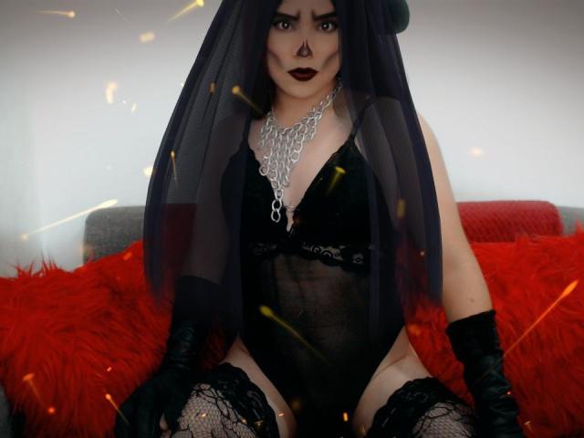 CamilaFetishDirty with dark makeup, black sheer veil, black lace lingerie, black gloves, and lace stockings, sitting on red fluffy pillows, showing her ass and black fishnet stockings.