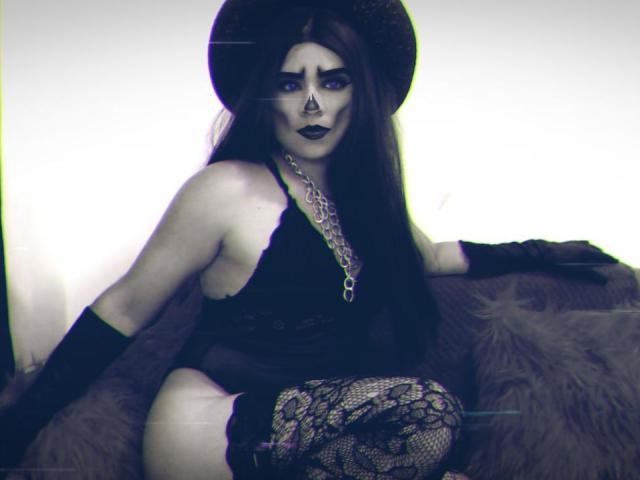 CamilaFetishDirty with long dark hair, wearing a black hat, lace bodysuit, and lace thigh-highs, sitting on a furry couch, black gloves, chain necklace, dark makeup, and a nose ring.