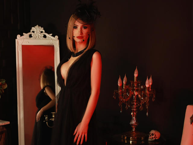 JuliaRichard con caschetto biondo, vestito nero, scollatura profonda, trucco sul viso e copricapo di piume, in piedi vicino a uno specchio, una candela rossa e un candelabro, con un teschio a destra..