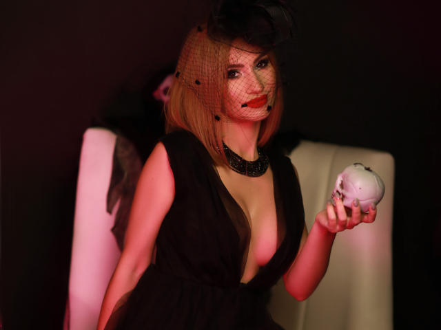 JuliaRichard con caschetto biondo, indossa un abito nero e un cappello a rete, tiene in mano un teschio bianco, scollatura profonda, collana nera, rossetto rosso, sexy, sfondo scuro, illuminazione rosa tenue..