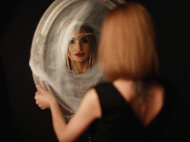 Julia Richard, avec ses cheveux blonds et son rouge à lèvres rouge, tenant un miroir rond, reflétant son visage, portant un haut noir sans manches, dans une image sombre, floue et avec un focus doux..