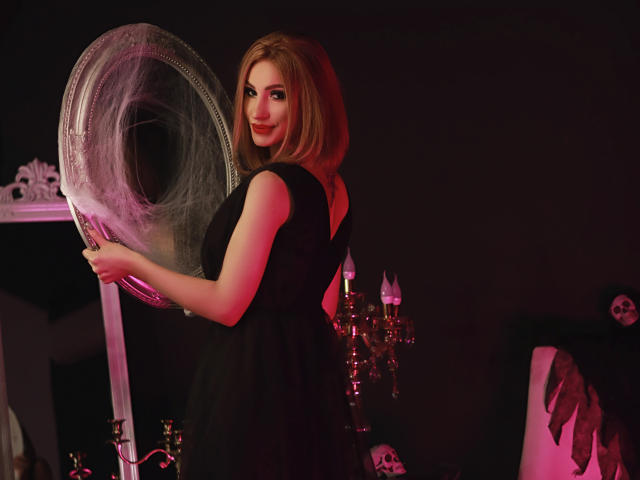 Julia Richard mit schulterlangem blondem Haar, in einem schwarzen Kleid, hält einen großen Spinnennetzrahmen, lächelt, rosa und rote Beleuchtung, Kerzenständer, Schädel im Hintergrund..