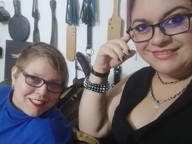 Two LatinoHardGirls with short brown hair and glasses, smiling at the camera. One wears a blue shirt, the other a black top with a necklace and spiked bracelet.