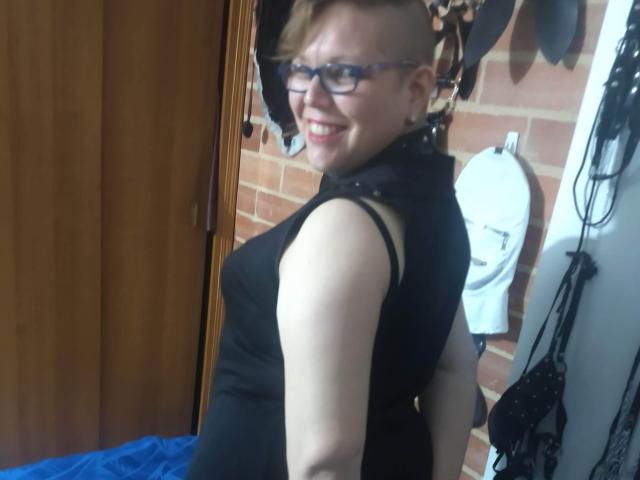LatinoHardGirls with short, buzzed side hair, glasses, black sleeveless top, smiling at camera, showing back, white urinal on brick wall, black bra hanging, wooden door, blue cloth.