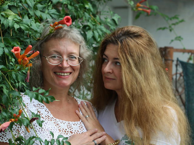 Duas mulheres, uma mais velha com cabelo grisalho, óculos e blusa branca de renda, e outra mais jovem com cabelo castanho comprido, em pé próximas, tocando as mãos, cercadas por flores laranjas..