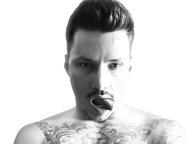 Fotografia in bianco e nero di TyagoDoll, senza camicia, con tatuaggi sul petto, capelli scuri corti, baffi, piercing al naso e la lingua fuori, che guarda leggermente di lato..