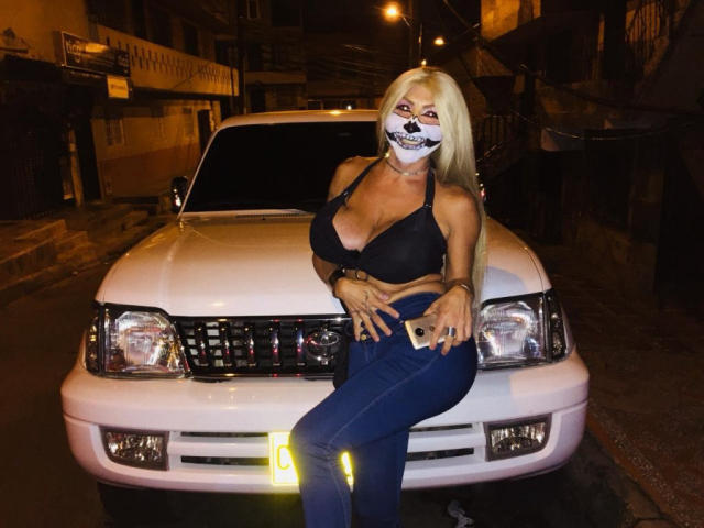 TsMonterCock avec des cheveux blonds, un soutien-gorge noir et un masque, assise sur une voiture blanche, en jean bleu, montrant des marques de bronzage, avec de grandes seins, tenant un téléphone, rue la nuit, phares de la voiture allumés..